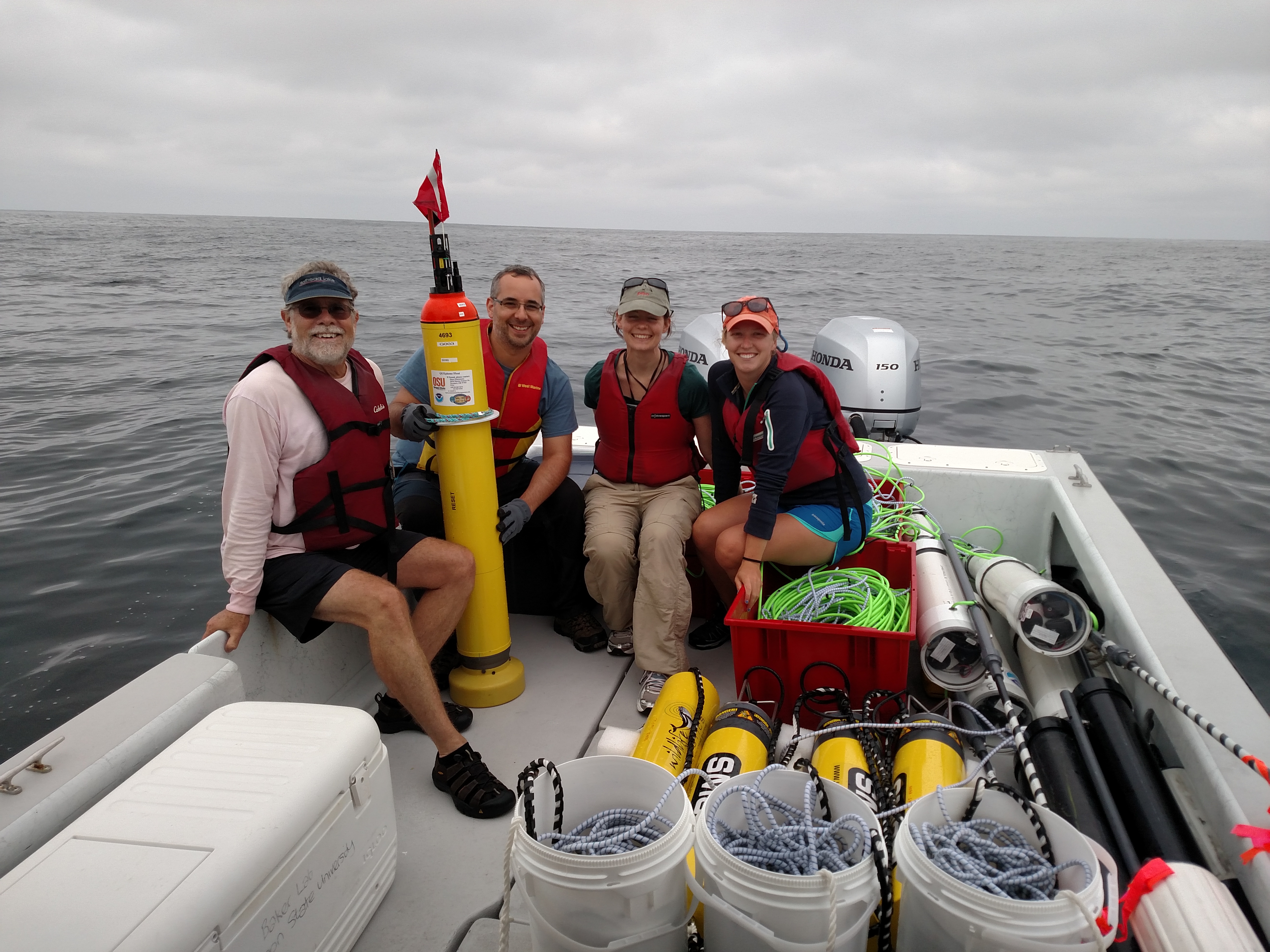 A group of four 
				smiling scientists  sitting on the back of a boat surrounded by several pieces of 
				scientific equipment. The skys are overcast and gray.