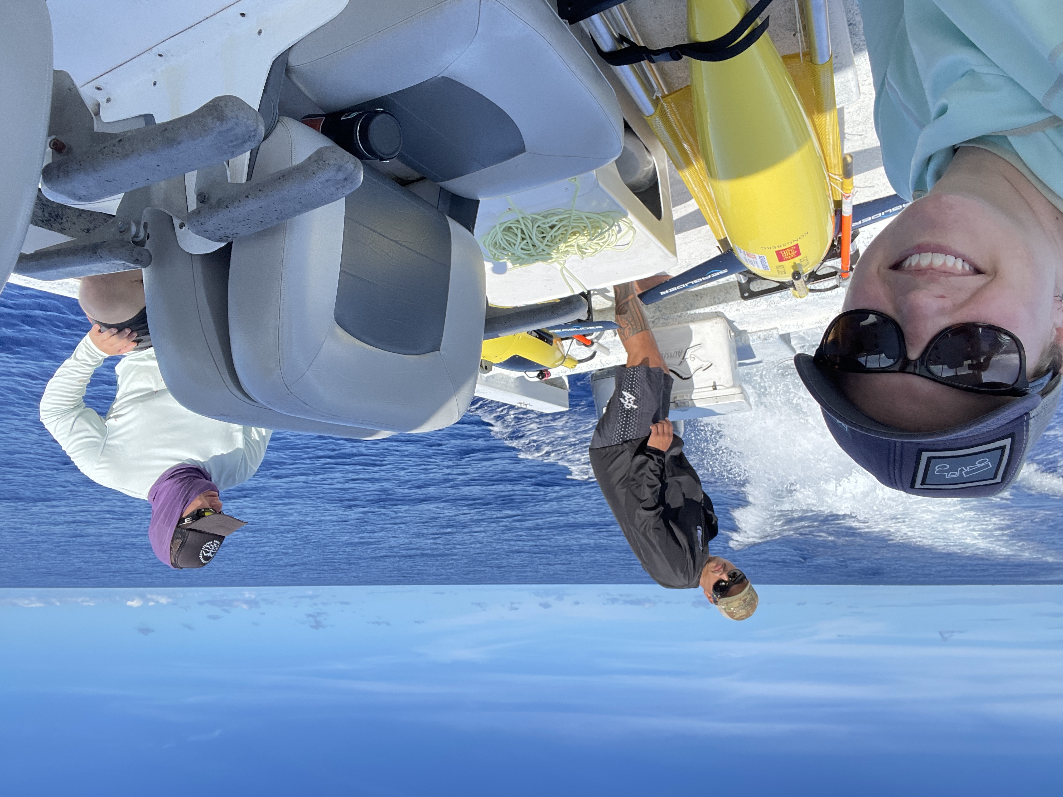 Three 
				smiling scientists on then back of a boat in sunshine on calm seas with two bright yellow 
				underwater gliders on the deck of the boat.