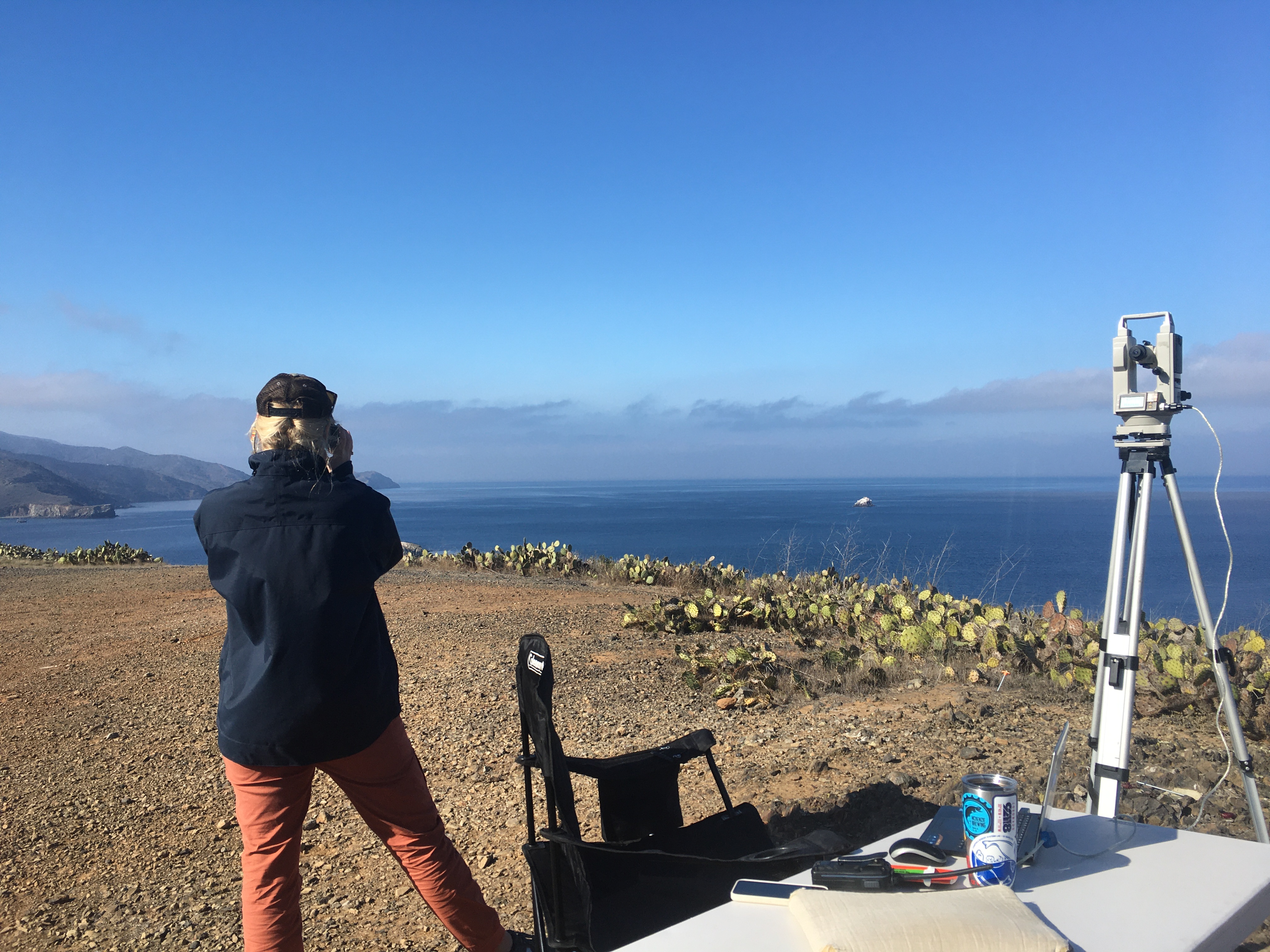 A person is standing on a cliff 
				overlooking the ocean. They are wearing a hat and black jacket standing next to a table with a 
				theodolite set up on their right.